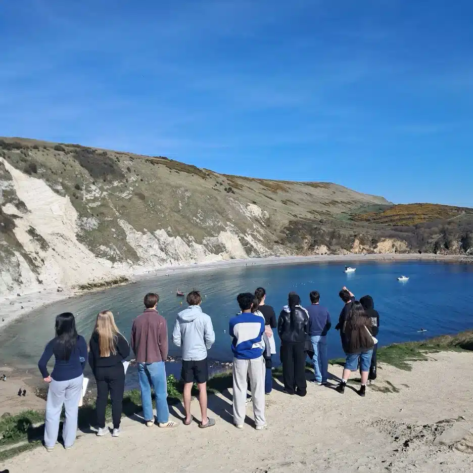 A group of people stand on a sandy cliff overlooking a scenic cove with clear blue water, white cliffs, and grassy hills under a bright blue sky. Some people are looking at the view, while others are talking.