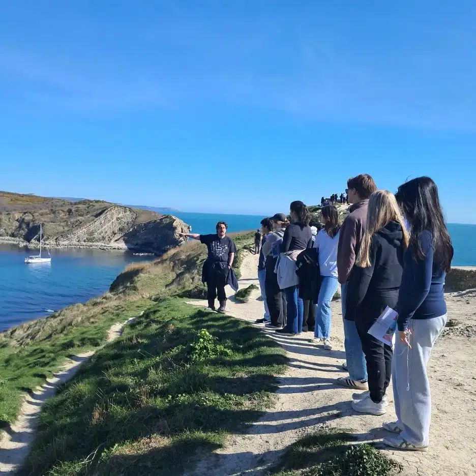 A group of people stand in a line on a coastal path overlooking the sea, listening to a person gesturing towards the water. A sailboat floats in a bay below, and the sky is clear and blue.
