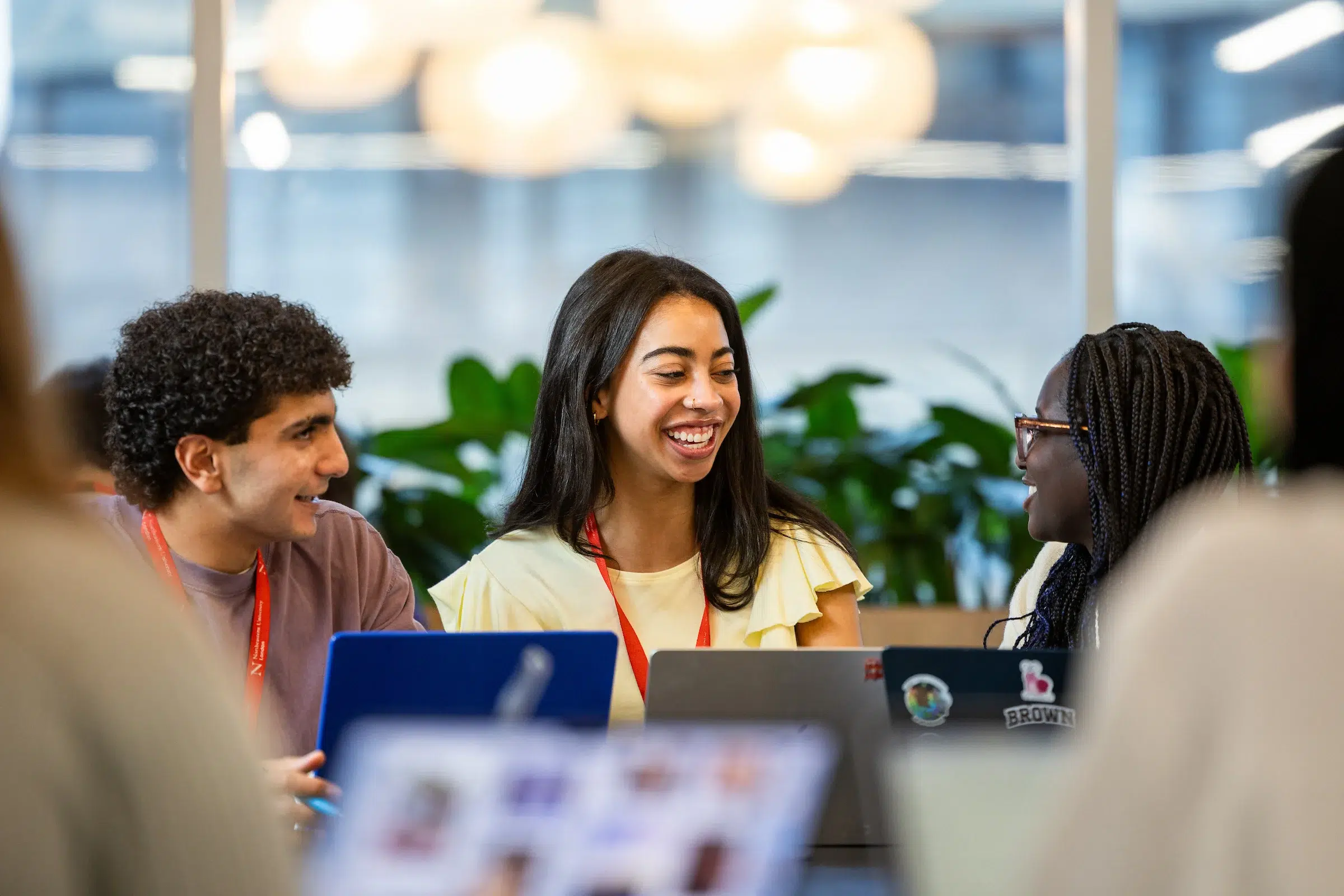 Three people sit at a table with laptops, smiling and talking in a bright, modern setting with green plants in the background—an engaging environment perfect for discussing Undergraduate Admissions.