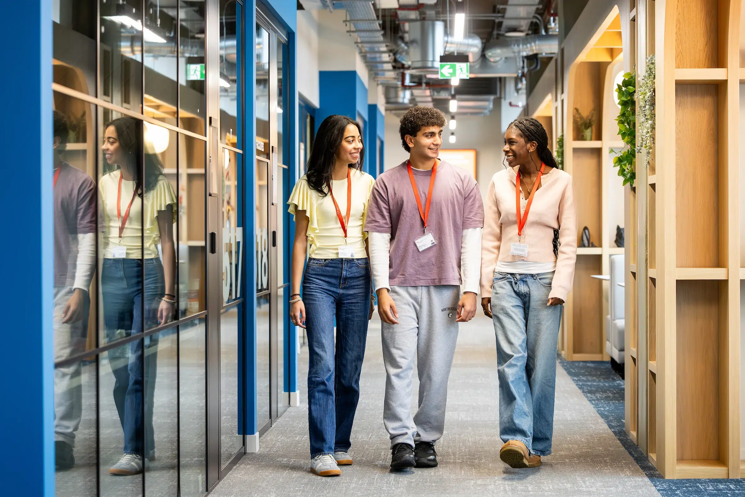 Undergraduate degrees. Three young adults wearing ID badges and casual clothes walk together down a modern office hallway, smiling and talking about their undergraduate degrees. There are glass walls on the left and wooden shelves with plants on the right.