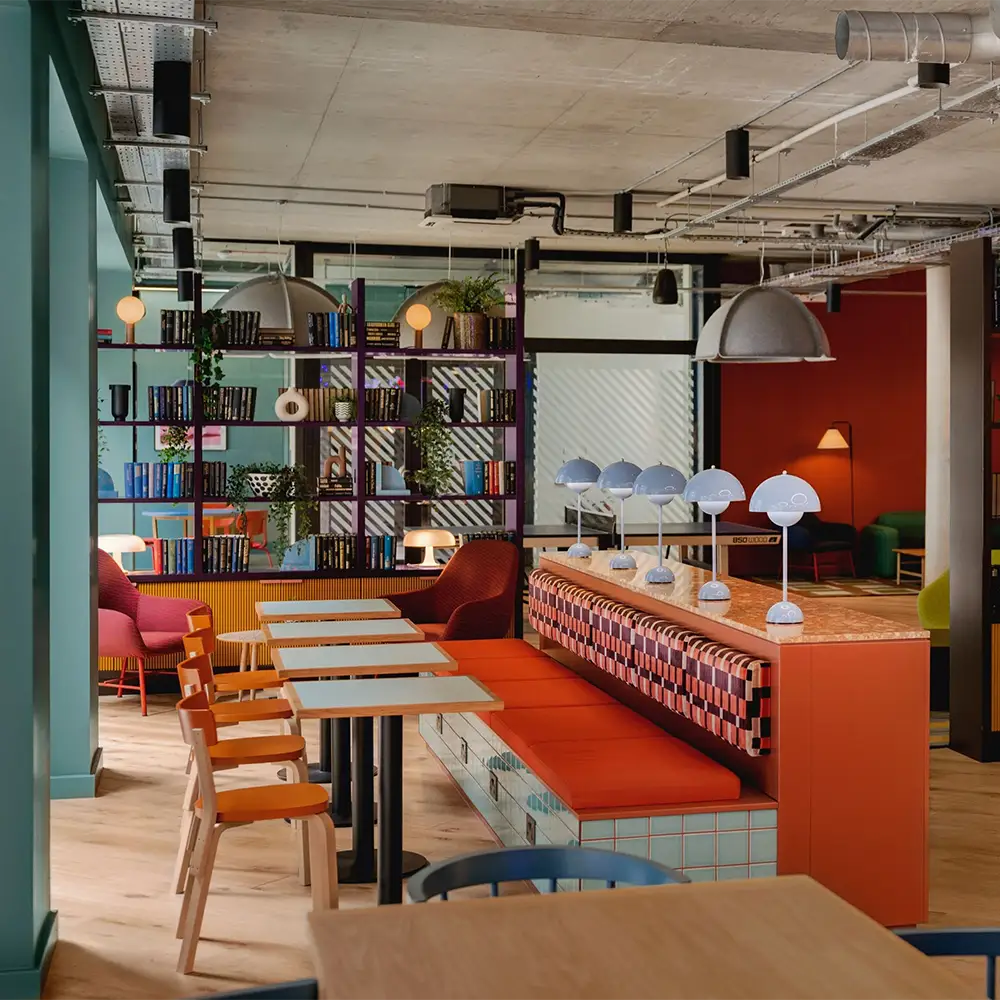 Scape Shoreditch. A modern, colorful workspace with wooden tables, orange and beige chairs, a cushioned bench, bookshelves, plants, and decorative lamps under exposed ceilings and industrial lighting offers comfortable accommodation for creative collaboration and productivity.
