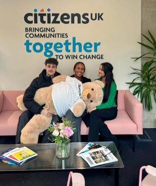 Three people sit on a pink sofa in front of a Citizens UK wall sign, holding a large teddy bear.