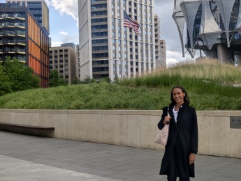 A smiling woman in a black coat and carrying a light-colored handbag stands in a plaza in front of high-rise buildings and an American flag.