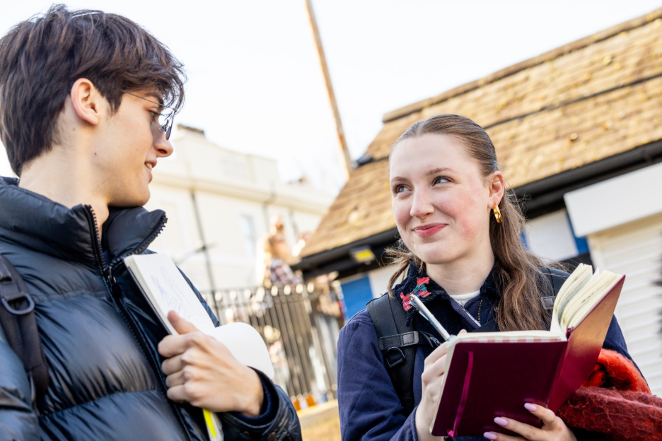 A young man holding a sketchbook talks to a young woman who is writing in a notebook.