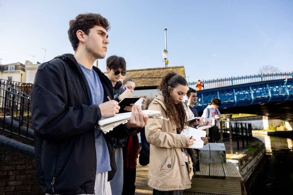 A group of students stand on a canal path, drawing in sketchbooks.